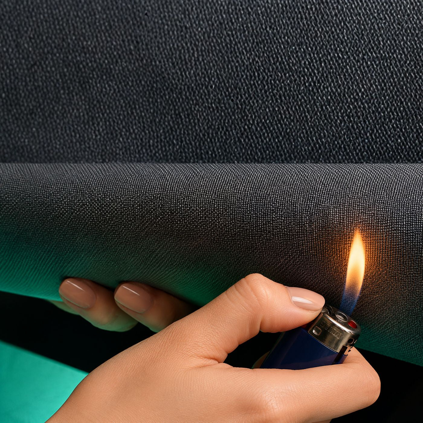 A close-up of a textured dark gray or black material being held by a hand, with a blue lighter lit and touching the surface to demonstrate heat resistance or flame resistance. Mass loaded vinyl